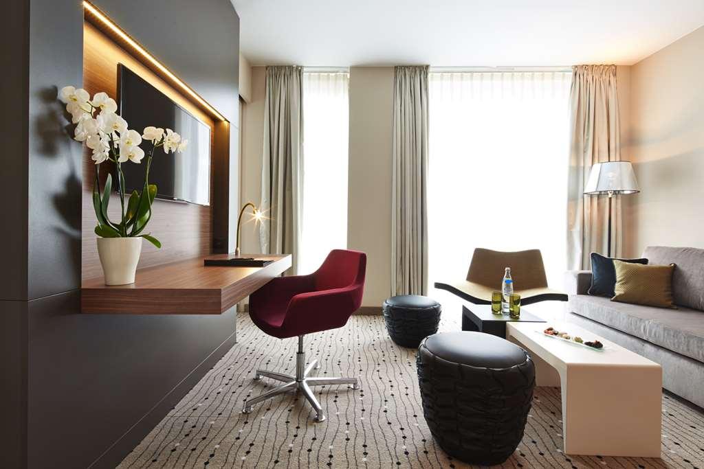 Steigenberger Hotel Am Kanzleramt Berlin Zimmer foto The photo depicts a modern and stylish living area. It features a sleek design with neutral-colored walls and large windows allowing natural light to fill the space. 

Key elements include:

- A wall-mounted television.
- A stylish red chair with a s