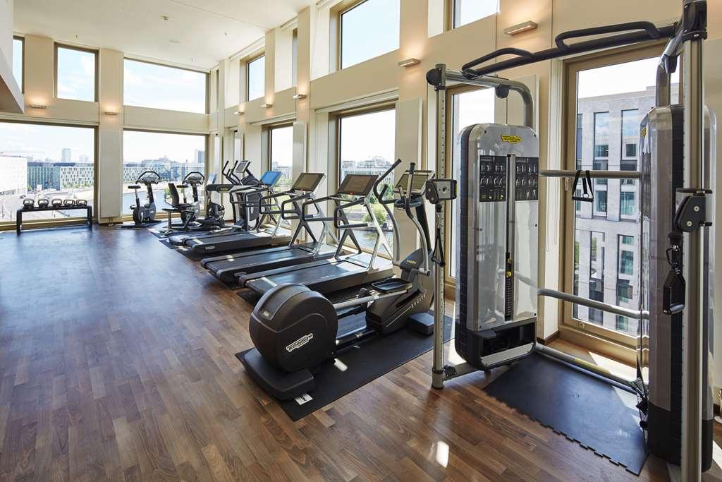 Steigenberger Hotel Am Kanzleramt Berlin Einrichtungen foto The photo shows a well-lit gym or fitness area with large windows allowing natural light to flow in. The gym is equipped with various exercise machines, including treadmills and elliptical trainers. There is also a multi-station strength training mac