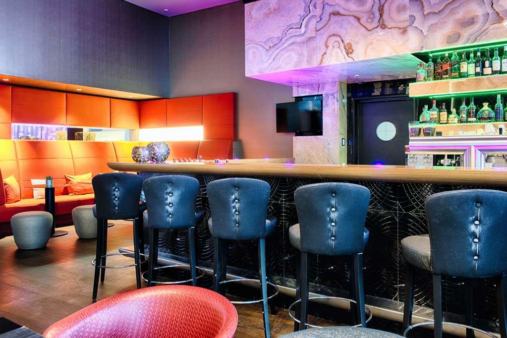 Steigenberger Hotel Am Kanzleramt Berlin Restaurant foto The photo shows a modern bar area featuring a long, sleek countertop with several black upholstered barstools arranged in front of it. The bar has decorative lighting that casts a colorful glow, enhancing the ambiance. Behind the bar, there is a sele
