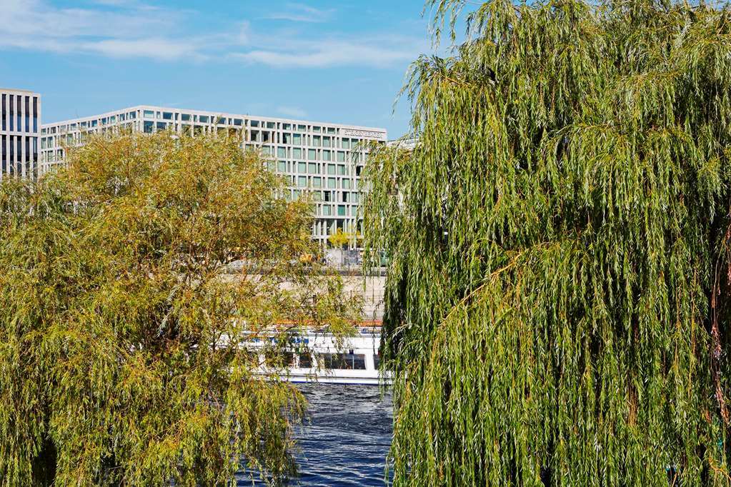 Steigenberger Hotel Am Kanzleramt Berlin Exterior foto The photo shows a view with lush, green willows in the foreground, characterized by their long, drooping branches. In the background, there is a modern building with large windows, suggesting a contemporary architectural style. The scene also include
