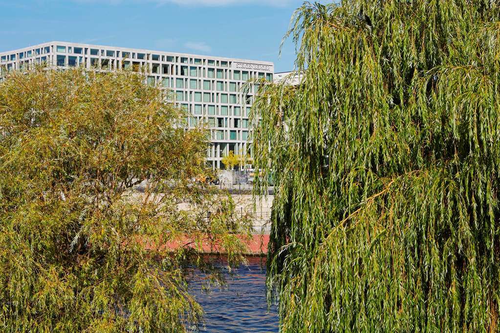 Steigenberger Hotel Am Kanzleramt Berlin Exterior foto The photo depicts a serene riverside scene with lush greenery. In the foreground, there are two willow trees with long, cascading branches that hang down toward the water. In the background, a modern building with a grid-like window design is visible