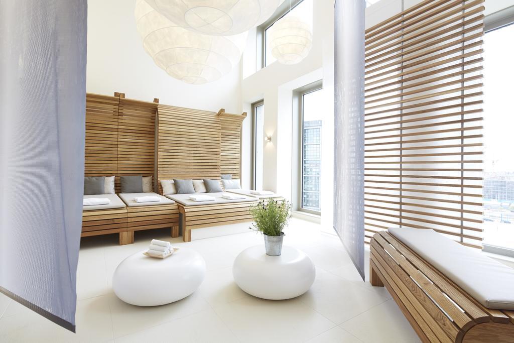 Steigenberger Hotel Am Kanzleramt Berlin Exterior foto The photo depicts a serene and modern interior space, likely a relaxation area or spa. It features wooden elements, including a slatted partition and lounge beds with soft cushions. There are round, white tables in the center, topped with a small pla