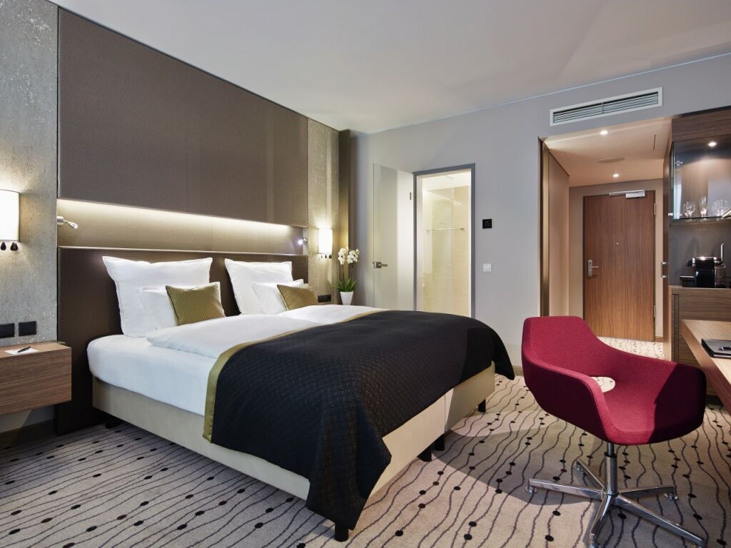 Steigenberger Hotel Am Kanzleramt Berlin Exterior foto The image shows a modern hotel room featuring a neatly made bed with white linens and a dark quilt cover. On the bed, there are two decorative pillows. The walls are a soft gray color, and there is subtle lighting above the headboard. To the side, th