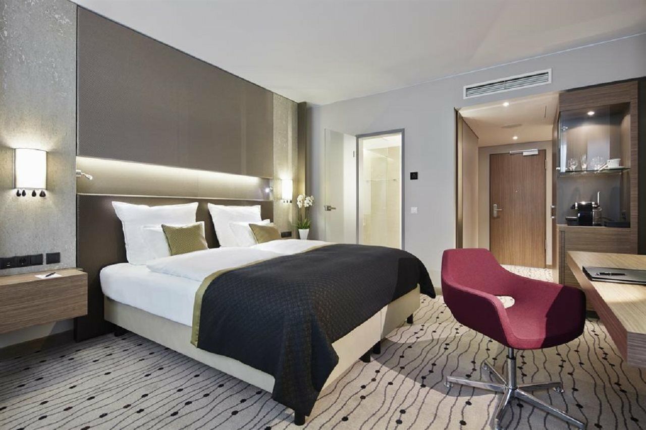 Steigenberger Hotel Am Kanzleramt Berlin Exterior foto The photo shows a modern hotel room featuring a double bed with white linens and a black throw blanket. There are two bedside lamps and a small table on one side. The room has a contemporary design with light gray walls and a patterned carpet. In the